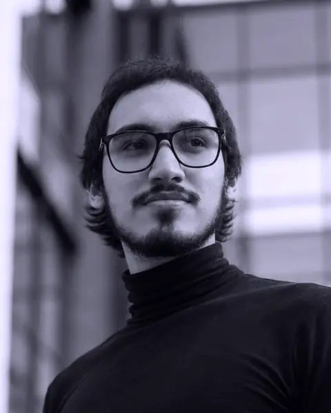 Portrait of Héctor Montero: in glasses and a black turtleneck, outdoors, between buildings.